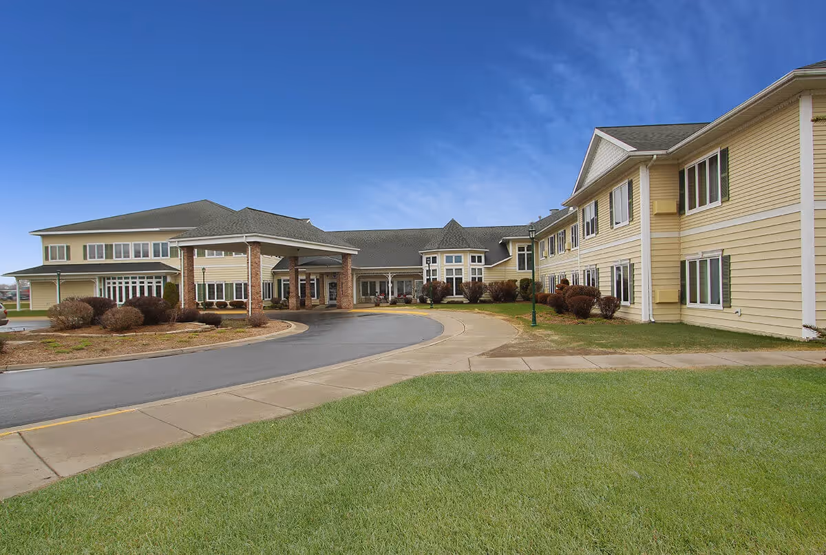 Front exterior of a large two-story yellow senior living facility with a covered entrance and circular driveway.