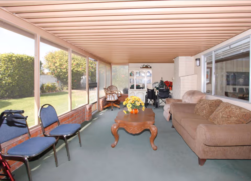 Senior living facility sunroom lounge with seating, coffee table, and garden view.