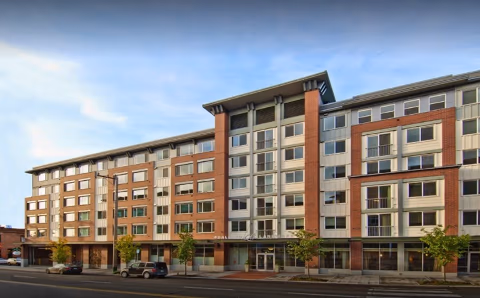 Exterior view of a multi-story senior living facility building with a modern design featuring large windows, brick and light-colored paneling, and a main entrance with glass doors. Several small trees and parked cars are visible in front of the building under a partly cloudy sky.