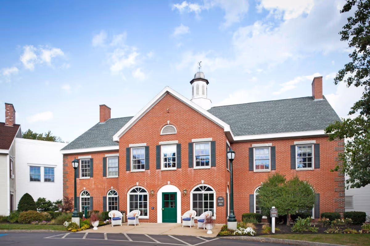 Two-story red-brick building with a green central door, arched windows, outdoor seating, and landscaped front under a blue sky.