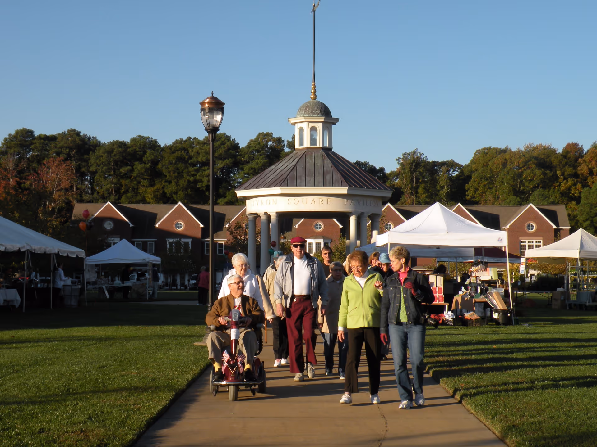 A group of elderly people walking and riding a mobility scooter on a paved path in a park-like area with green grass and tents set up for an event. In the background, there is a pavilion with columns and a cupola, and brick buildings surrounded by trees under a clear blue sky.