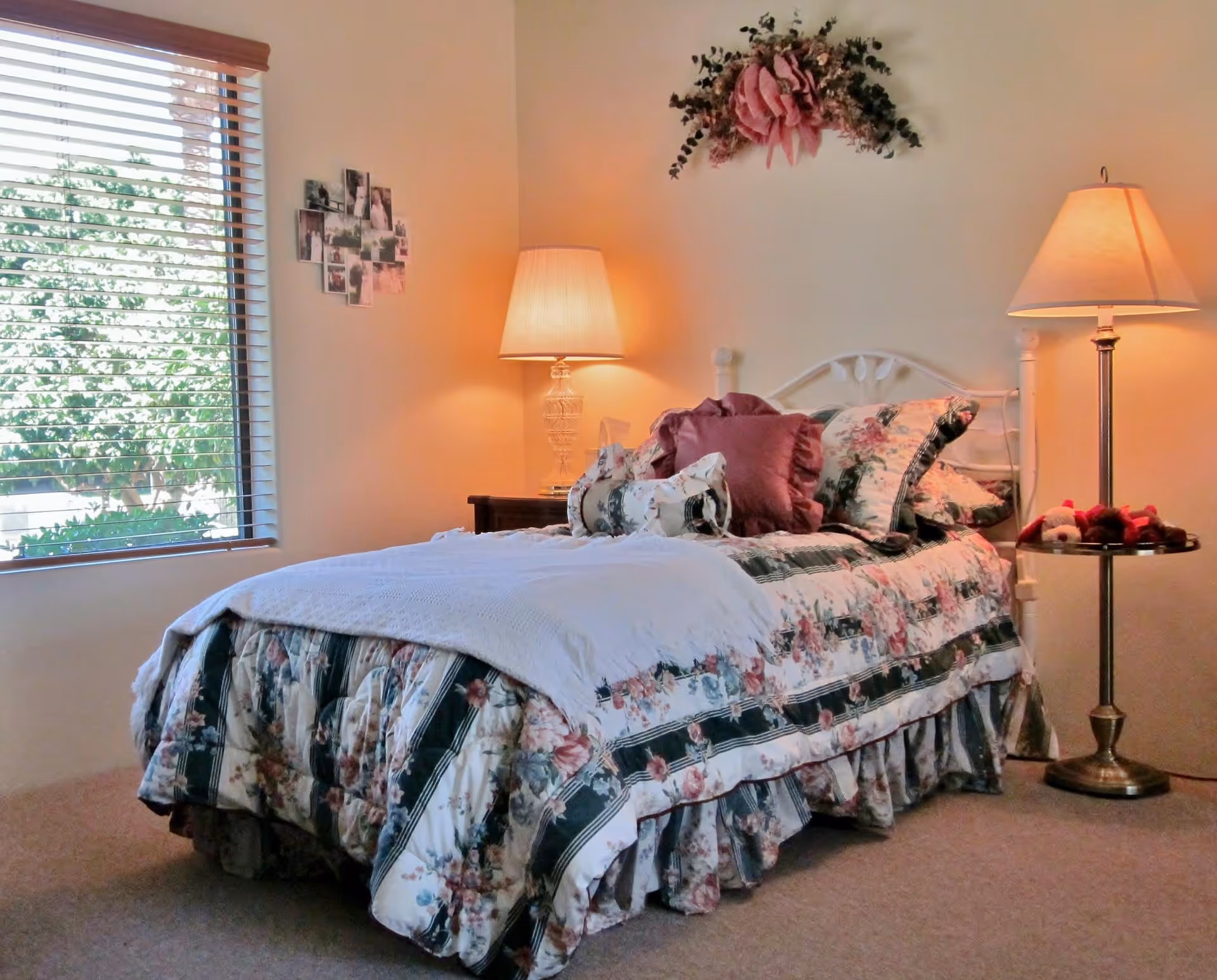 A cozy bedroom with a floral patterned bedspread and multiple pillows on a white metal bed frame. There are two lamps on either side of the bed, one on a wooden nightstand and the other on a small round table with some stuffed animals. A window with wooden blinds lets in natural light, and a floral wall decoration hangs above the bed.
