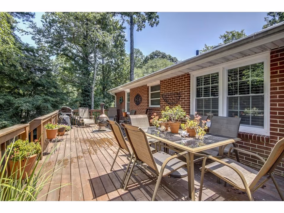 A wooden outdoor deck with patio table, chairs, potted plants, and trees beside a brick home