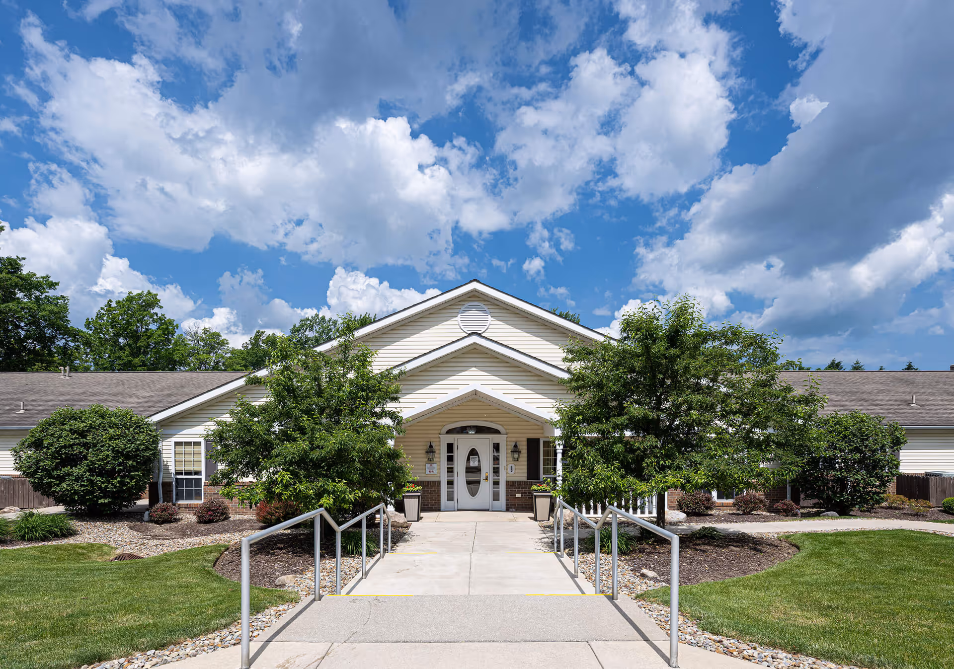 Front exterior view of a single-story building with a peaked roof and white siding, surrounded by green trees and shrubs under a partly cloudy blue sky. A concrete walkway with handrails leads to the entrance door.