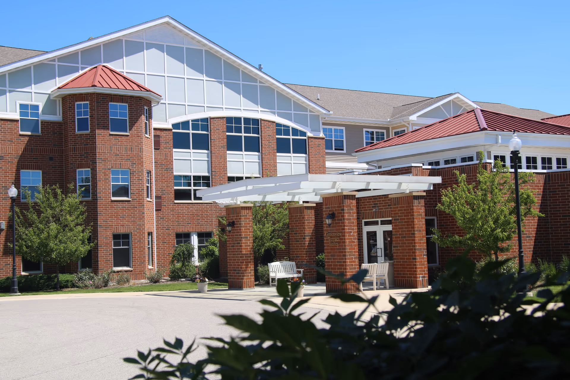 Exterior view of a senior living facility building with red brick walls, large windows, and a covered entrance supported by brick pillars. There are benches under the covered area and greenery including trees and bushes around the building. The sky is clear and blue.