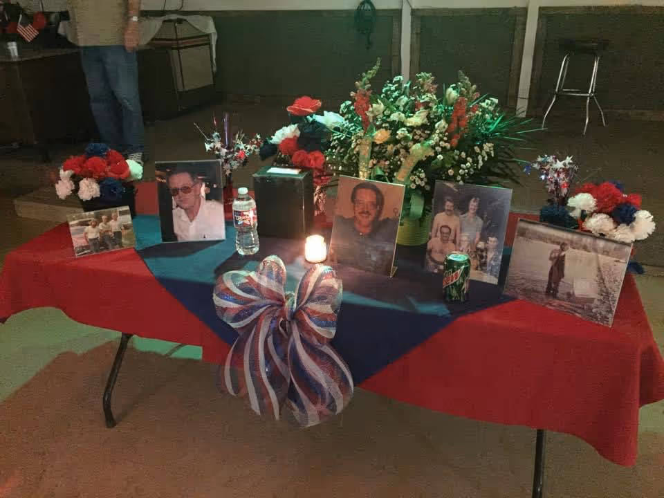 Memorial table with framed photos, flowers, candles, and a bow on a red-blue cloth.