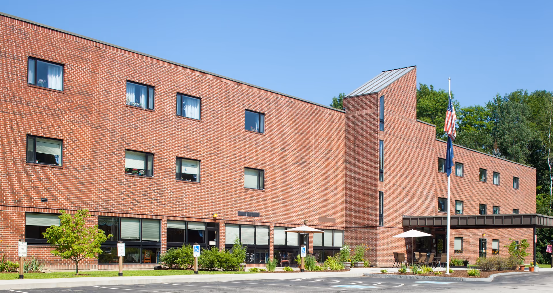 Exterior view of a three-story brick building with multiple windows, a flagpole with an American flag, and a small outdoor seating area with umbrellas. The building is surrounded by greenery and a parking lot with handicap parking spaces.