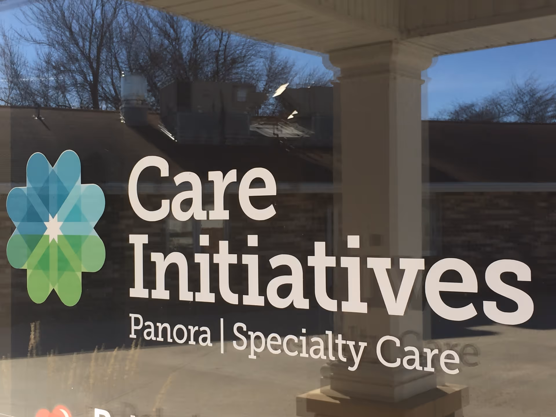 Window decal displaying the logo and name of Care Initiatives Panora Specialty Care, with a geometric flower-like design in blue and green colors, reflected against an outdoor scene with trees and a building.