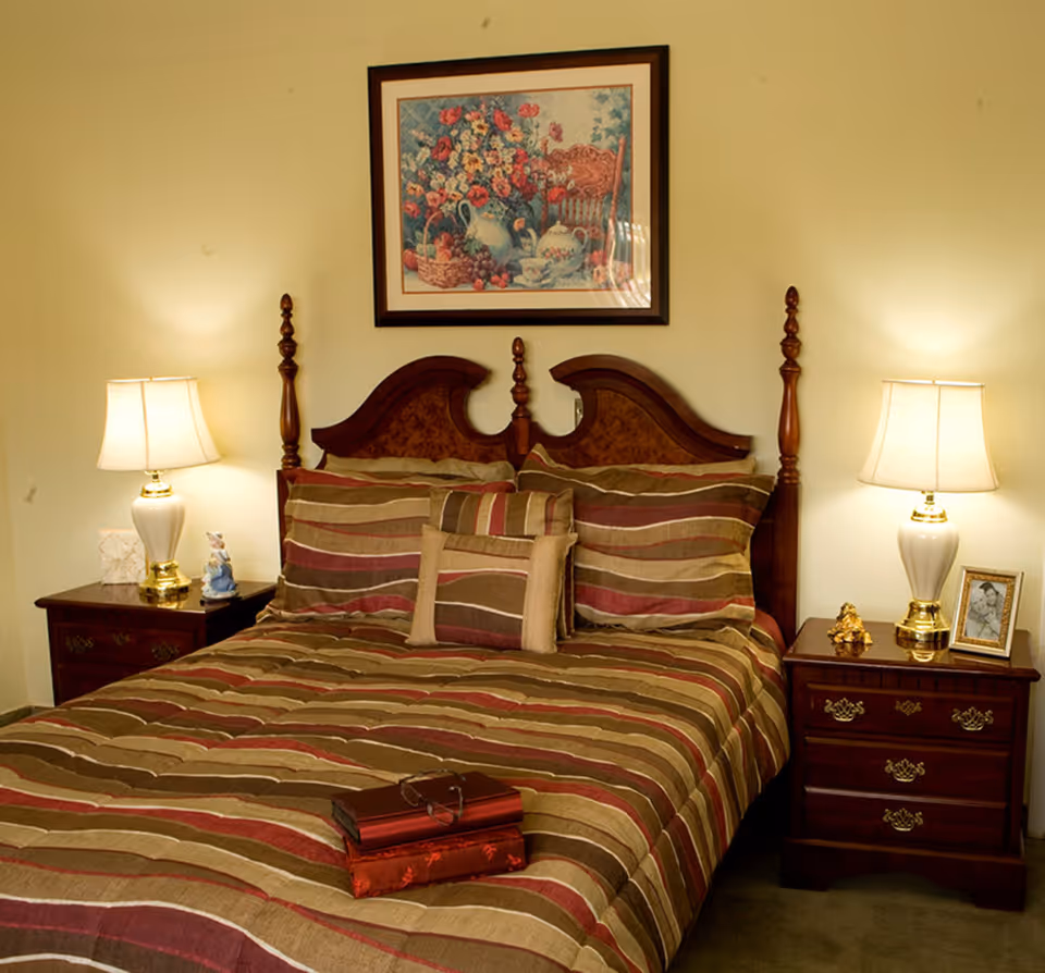 A neatly made bed with a wooden headboard and striped bedding in shades of brown, beige, and red. There are two matching pillows and a smaller decorative pillow. On the bed are two closed books with a pair of glasses resting on top. On either side of the bed are wooden nightstands with brass handles, each holding a white lamp with a beige lampshade. The left nightstand also has a small figurine and a tissue box, while the right nightstand has a small golden decorative item and a framed black-and-white photo. Above the bed hangs a framed painting of a floral arrangement with a basket of fruit.