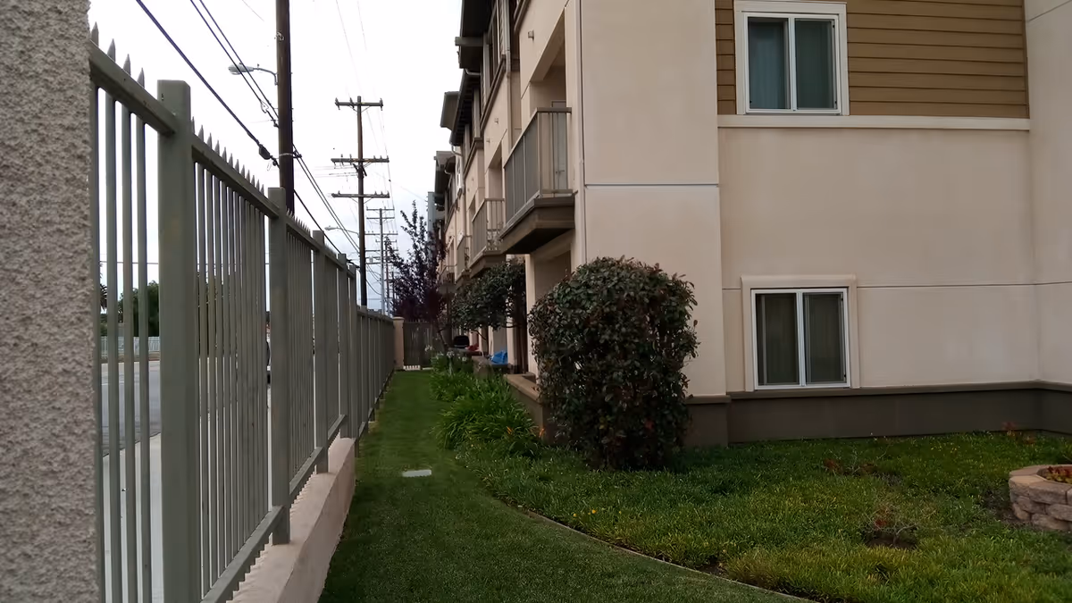 Exterior view of a residential building with a side lawn, bushes, and a metal fence.