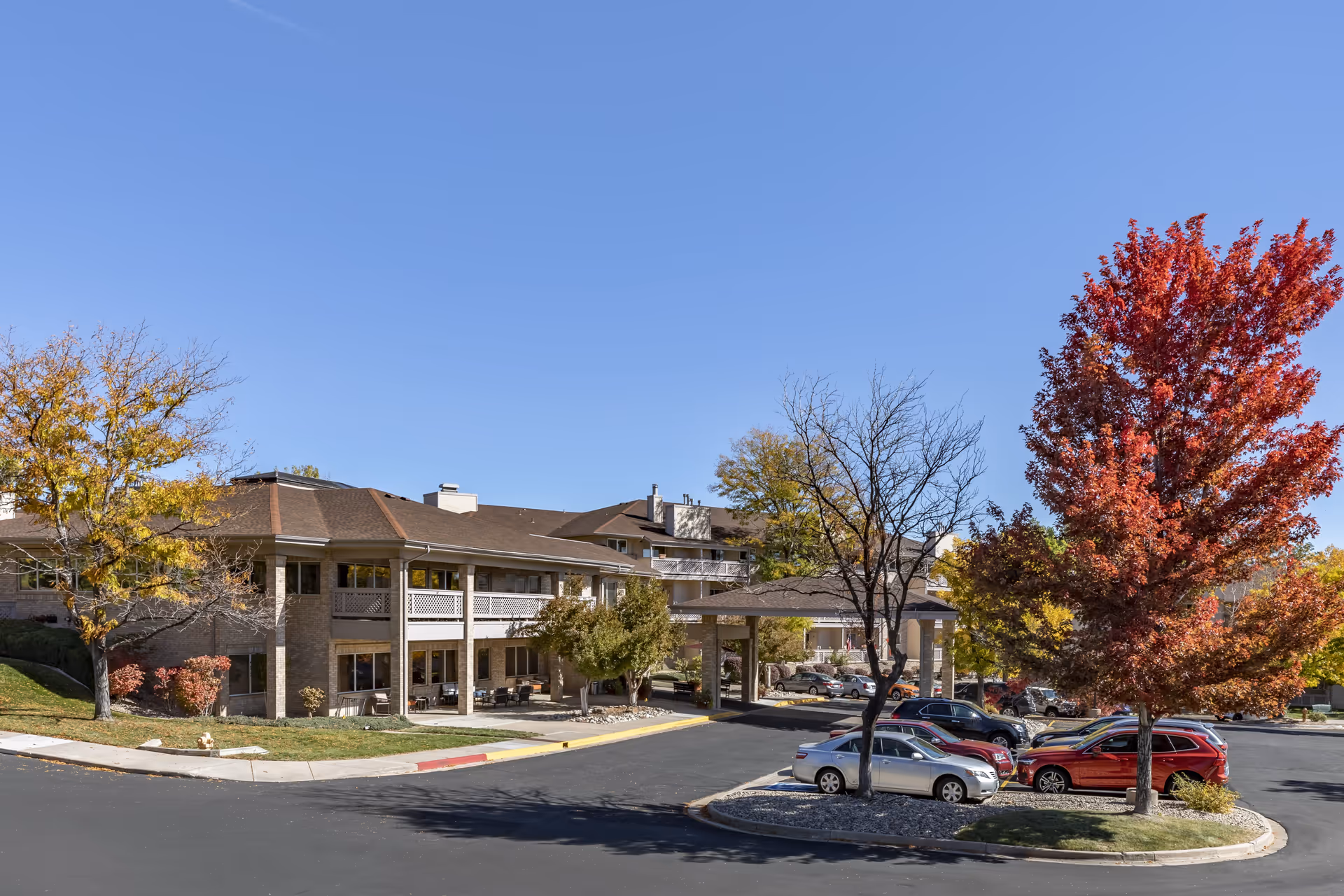 Exterior view of Brookdale Meridian Westland senior living facility showing a two-story building with a covered entrance, surrounded by trees with autumn foliage and a parking lot with several cars under a clear blue sky.