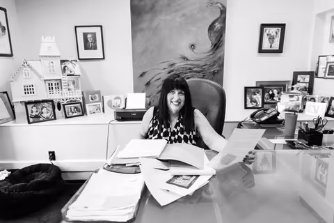 Smiling woman sits at a glass desk surrounded by papers and photos in an office.