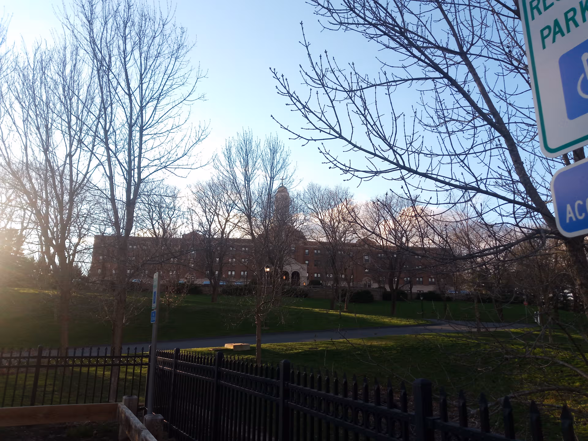 Front view of a brick senior living facility beyond leafless trees and a fence.