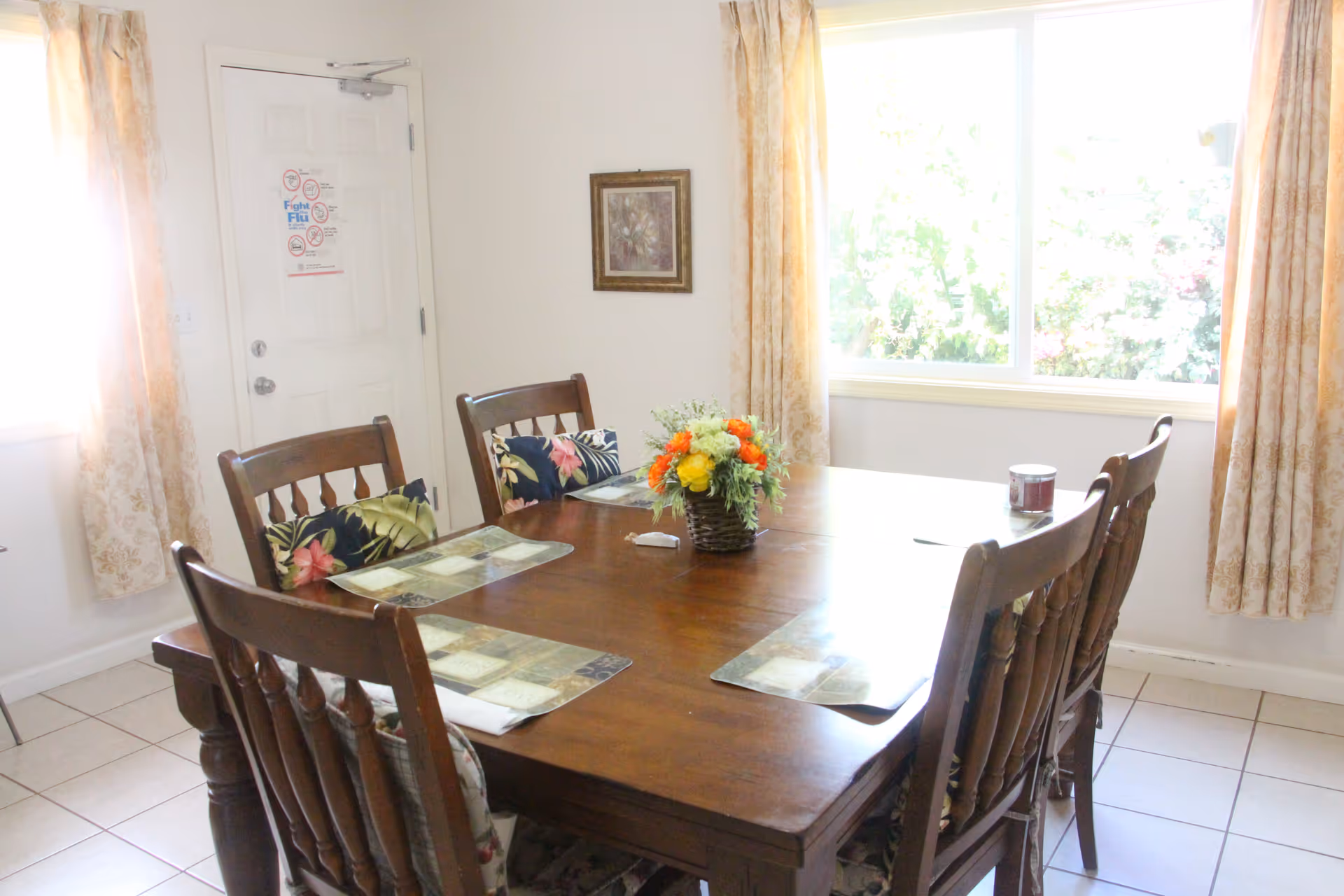 Wooden dining table with six chairs and a flower centerpiece in a bright dining room.