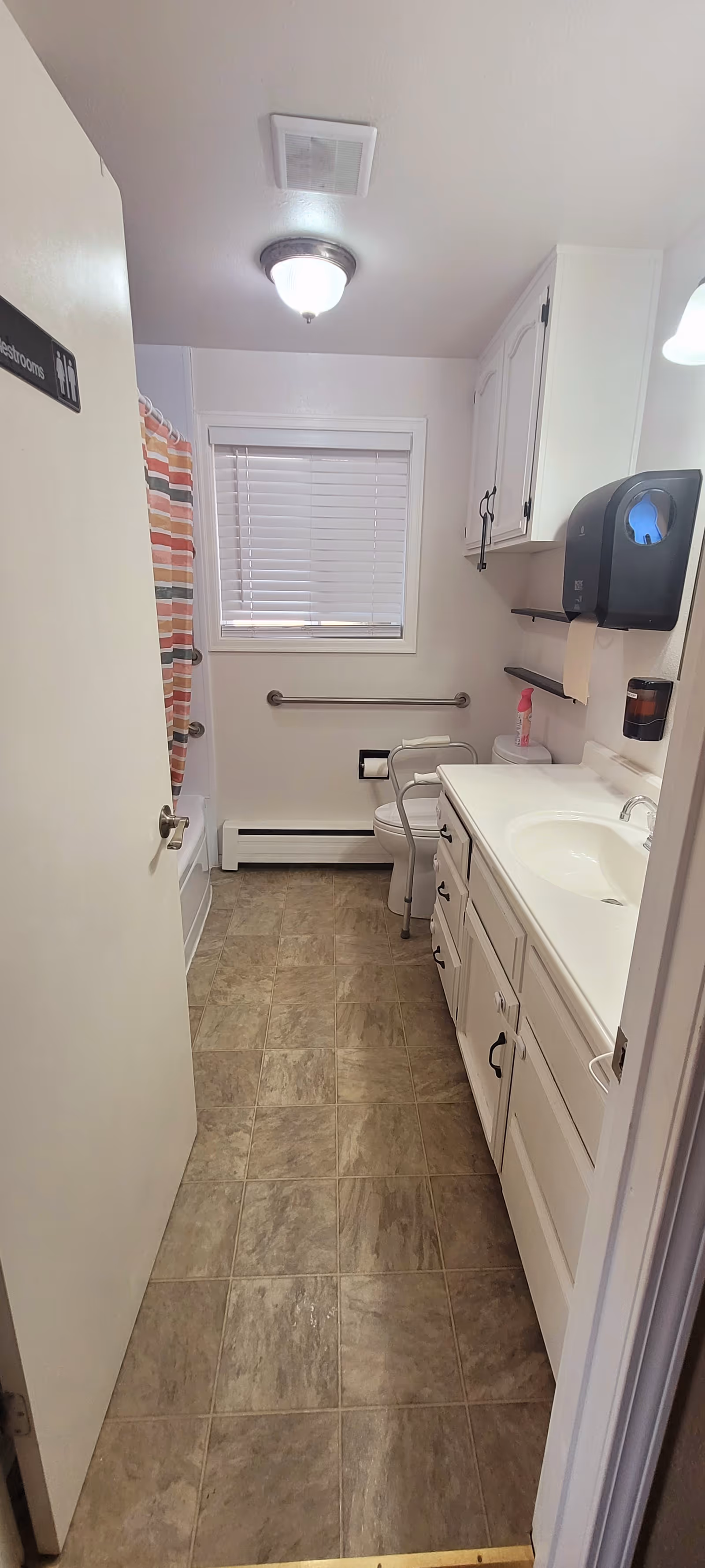 Small accessible bathroom in a senior facility with sink vanity, toilet, grab bars, and shower.