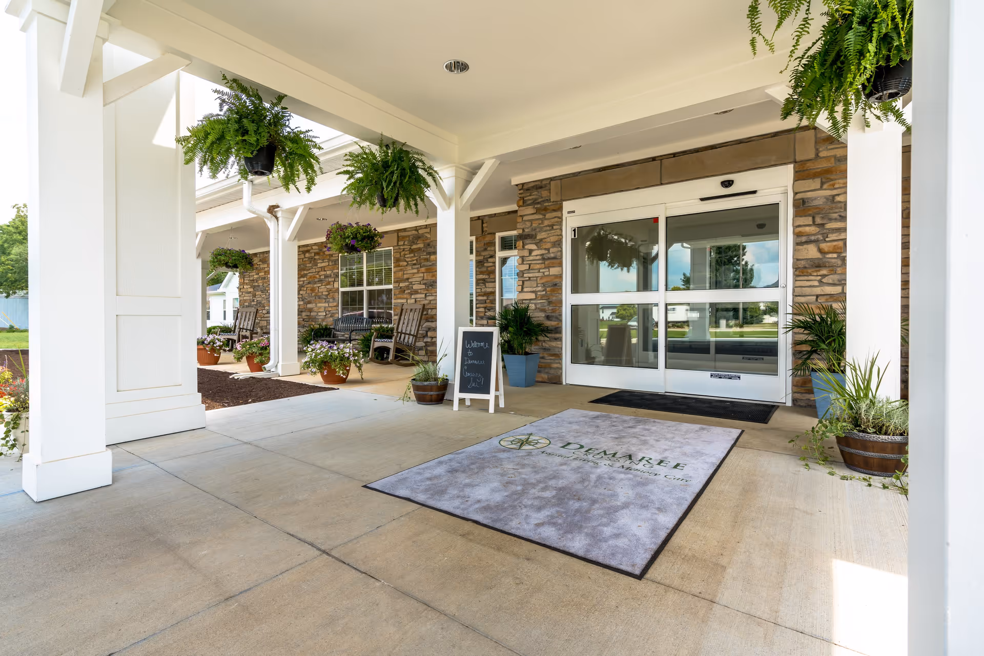 Covered entrance area of Demaree Crossing with hanging ferns, potted plants, rocking chairs, and a welcome sign near automatic glass doors. The building exterior features stone walls and white pillars.