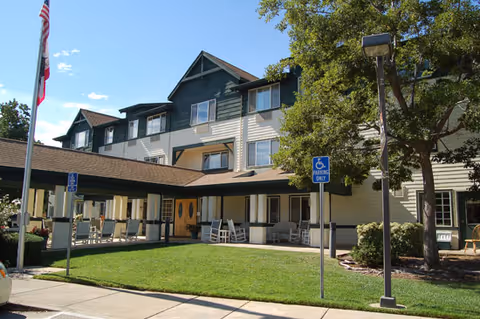 Front entrance of a multi-story senior living building with a covered driveway, porch rocking chairs, flags, and a small lawn.