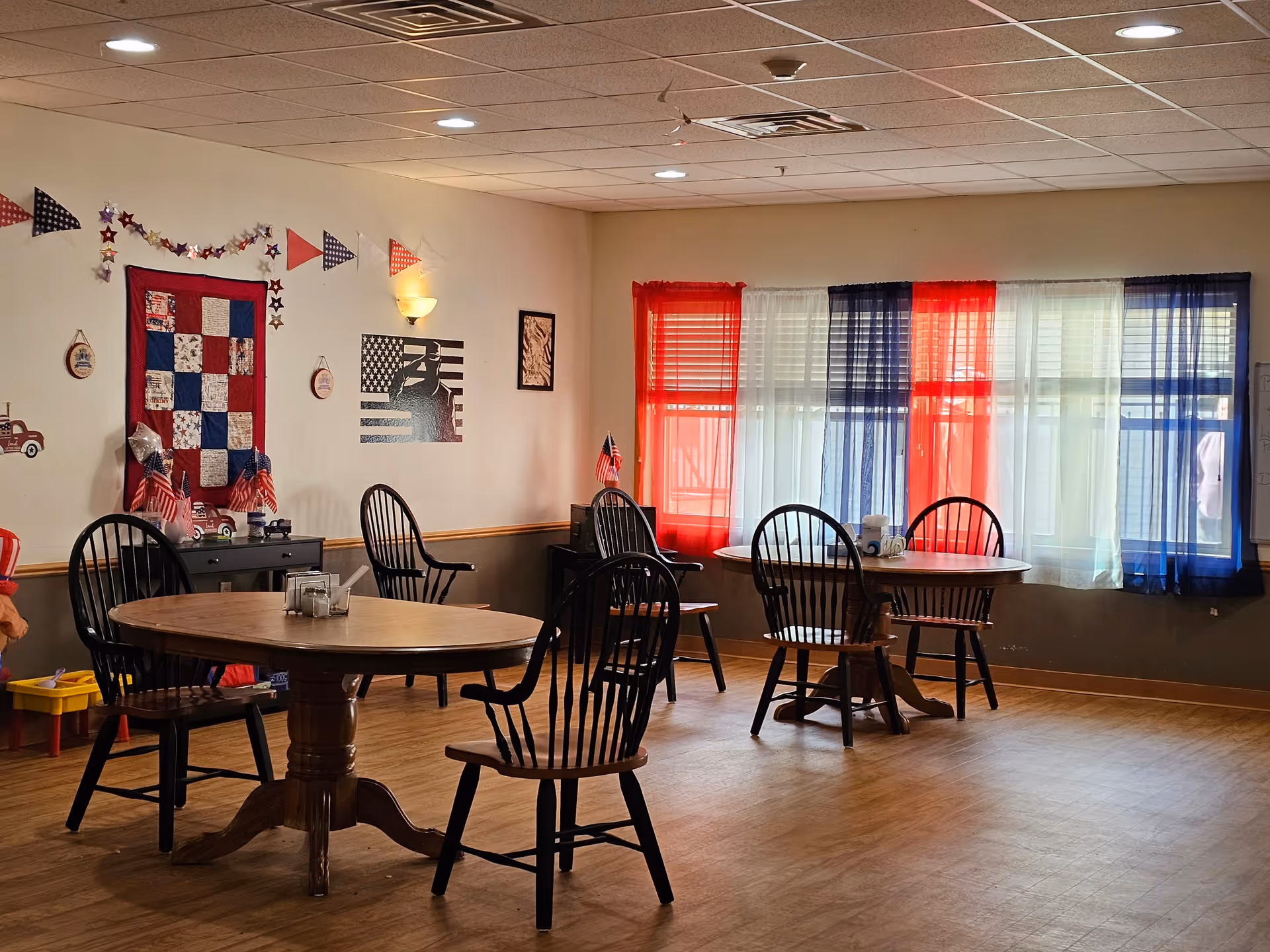 A dining room with wooden round tables and black wooden chairs. The room is decorated with patriotic-themed items including red, white, and blue curtains, a quilt on the wall, small American flags, and wall art featuring stars and stripes. The floor is wooden, and the ceiling has recessed lighting.