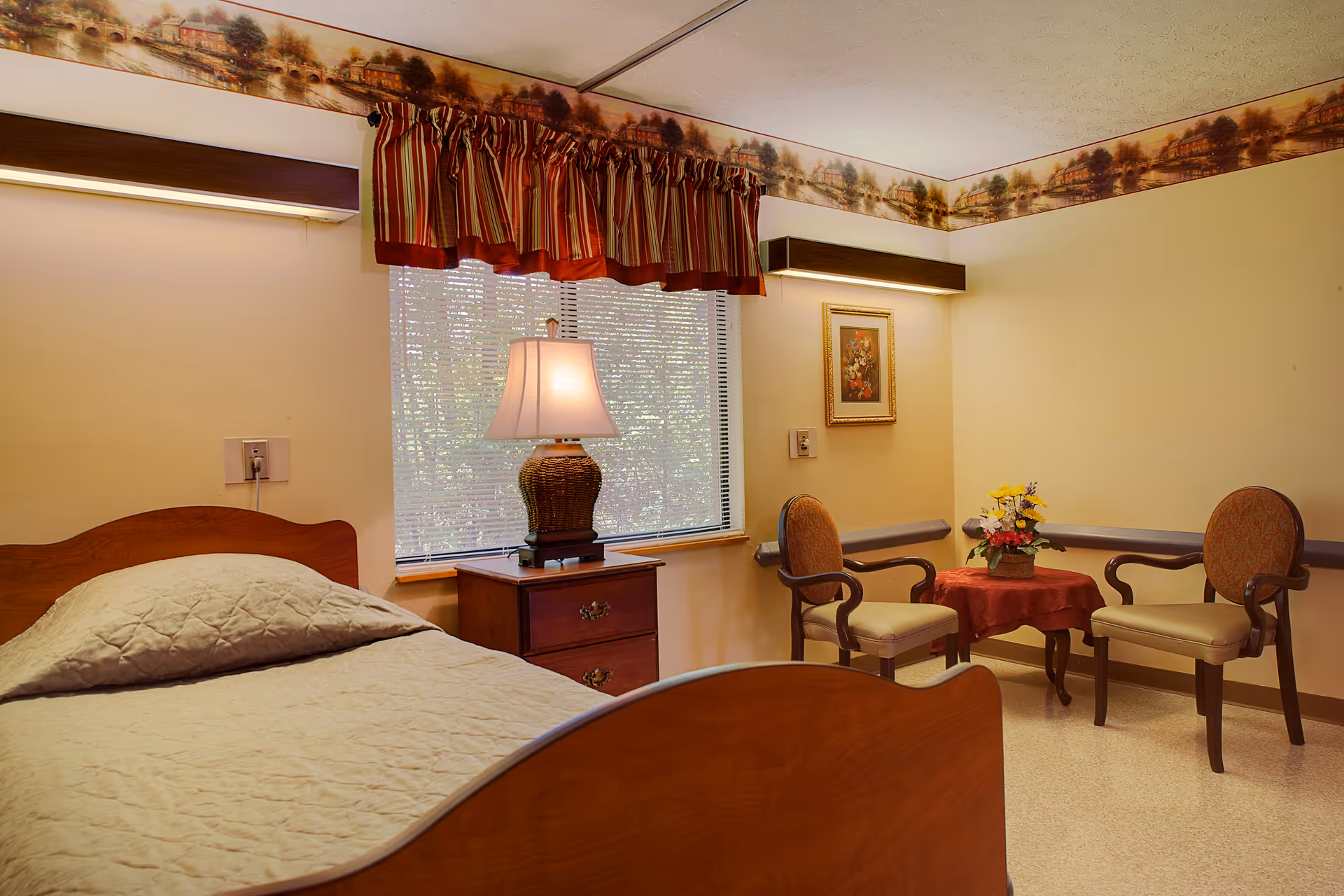 A cozy bedroom in a senior living facility featuring a single wooden bed with a beige quilt, a wooden nightstand with a lamp, two upholstered armchairs around a small table with a red tablecloth and a flower arrangement, a window with striped valance curtains, and a framed floral picture on the wall.