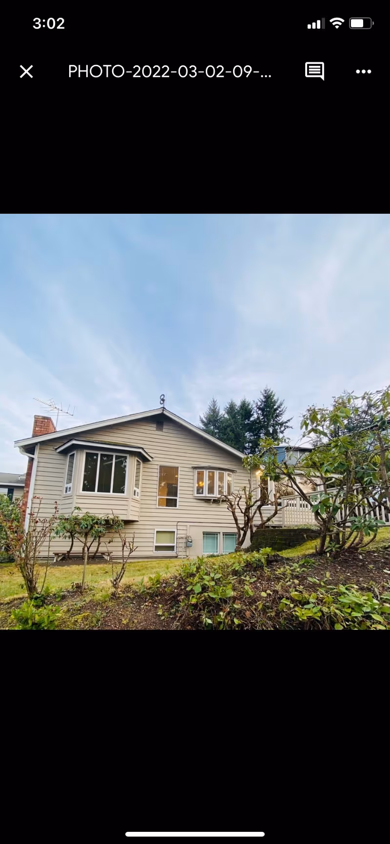 Front exterior of a single-story beige house with bay windows, shrubs, and a small landscaped yard under a blue sky.