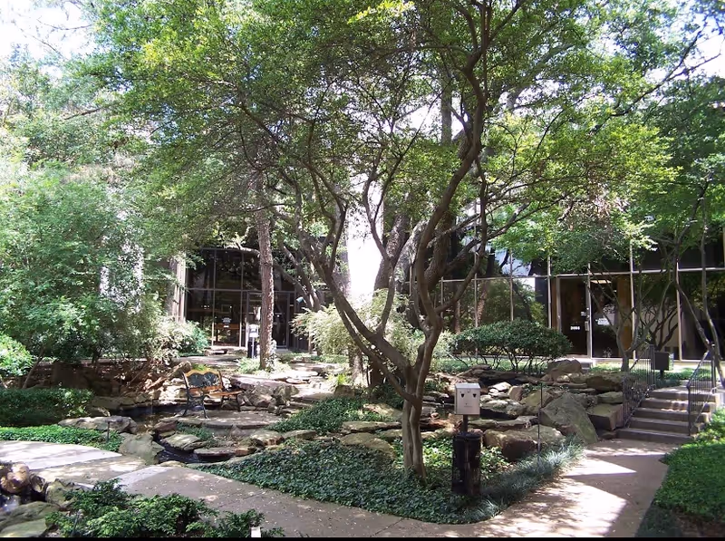 Outdoor courtyard with trees, benches, stone paths, and a glass-front building.