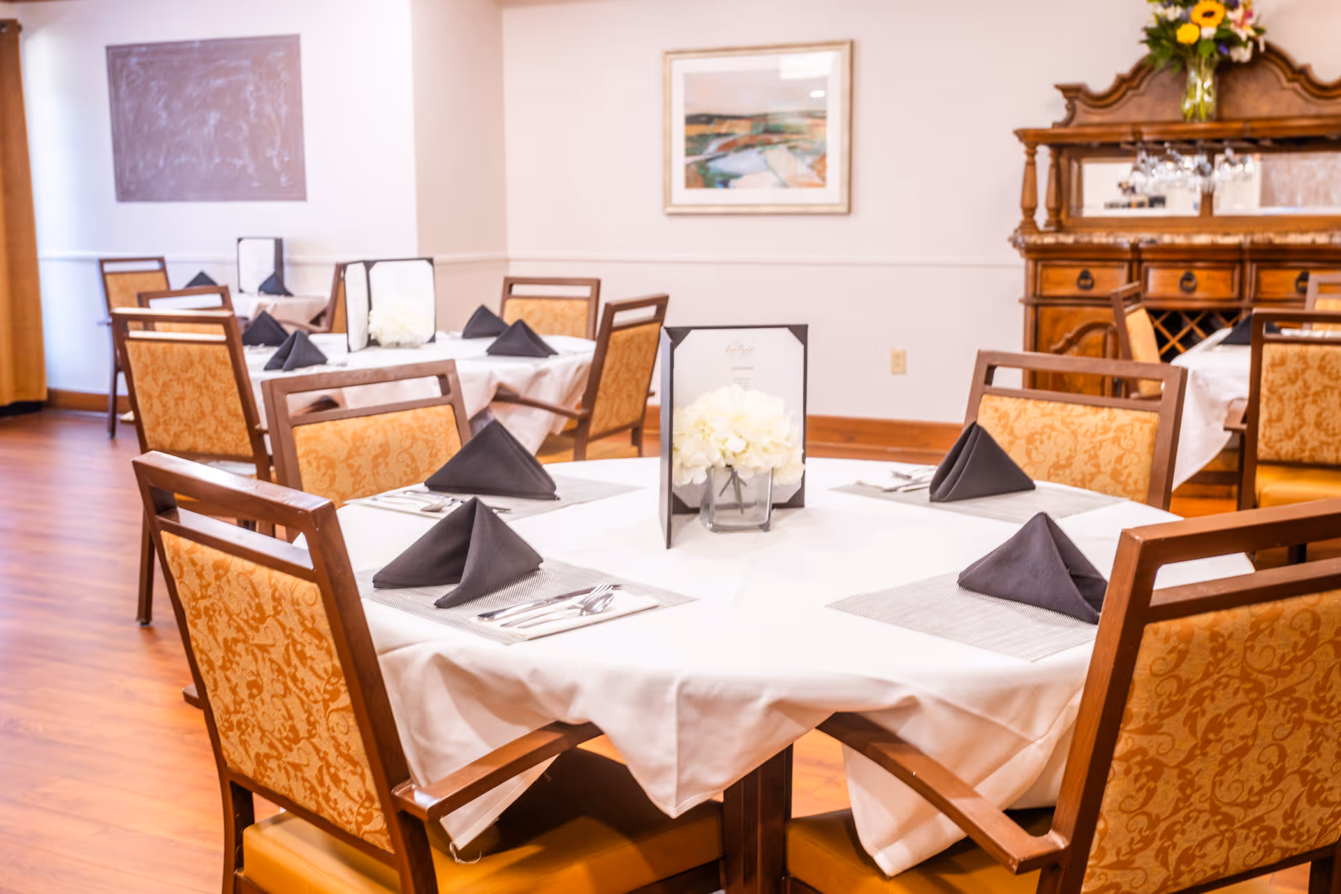 Round tables set for dinner with white tablecloths, black napkins, and floral centerpiece in dining room.
