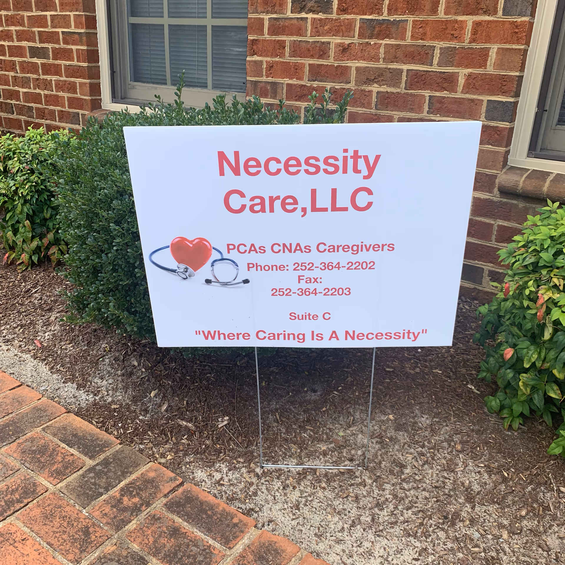 White sign with red text reading Necessity Care, LLC outside a brick building.