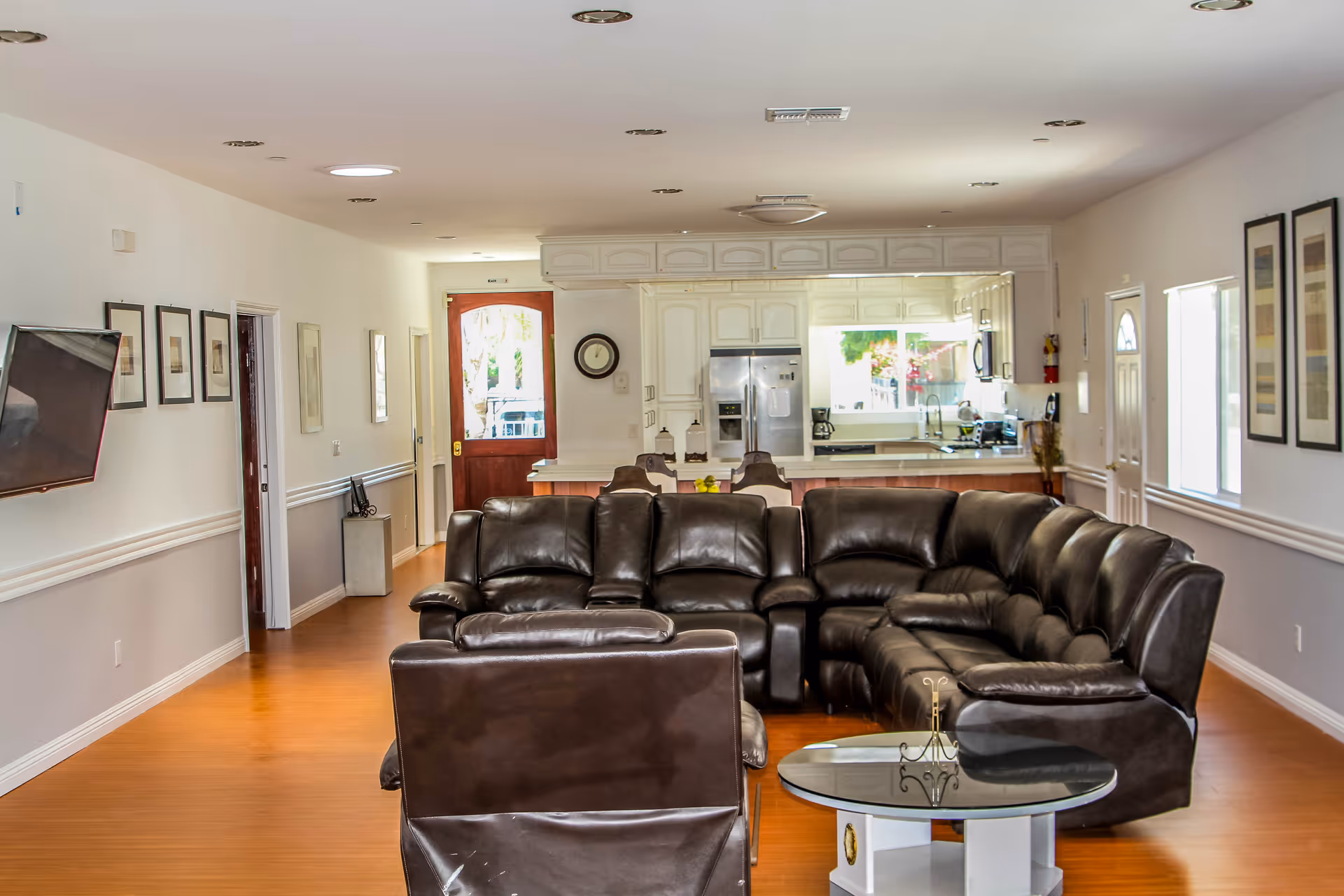 Interior view of a senior living facility common area featuring a large dark brown leather sectional sofa and a matching chair in the foreground. Behind the seating area is a kitchen with white cabinetry, stainless steel refrigerator, and a window letting in natural light. The walls are decorated with framed artwork, and the floor is a warm wood tone. A wall-mounted TV is visible on the left side.