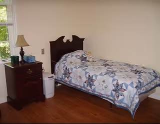 Small bedroom with a single bed covered by a patterned quilt, a wooden nightstand with a lamp, and a window.