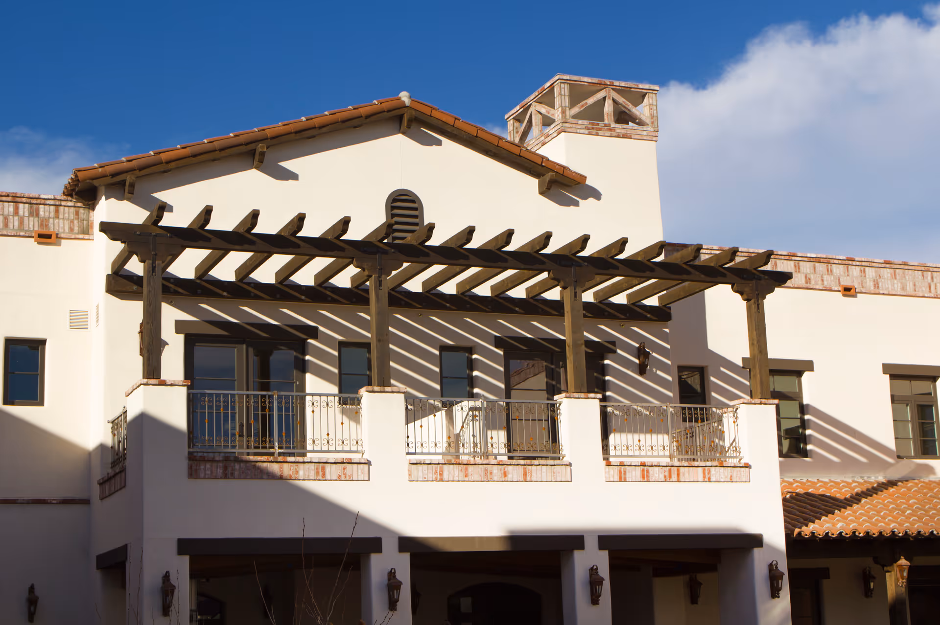 Exterior view of a building with a white facade and a wooden pergola on the balcony. The building has a tiled roof and several windows, with clear blue sky in the background.