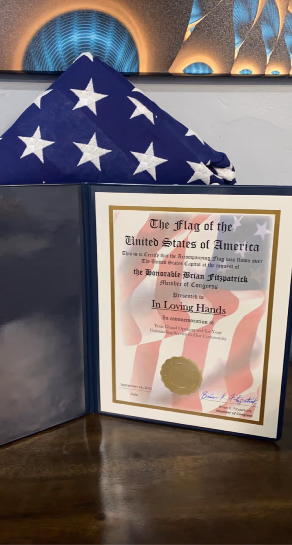 Framed certificate titled In Loving Hands beside a folded American flag on a desk.