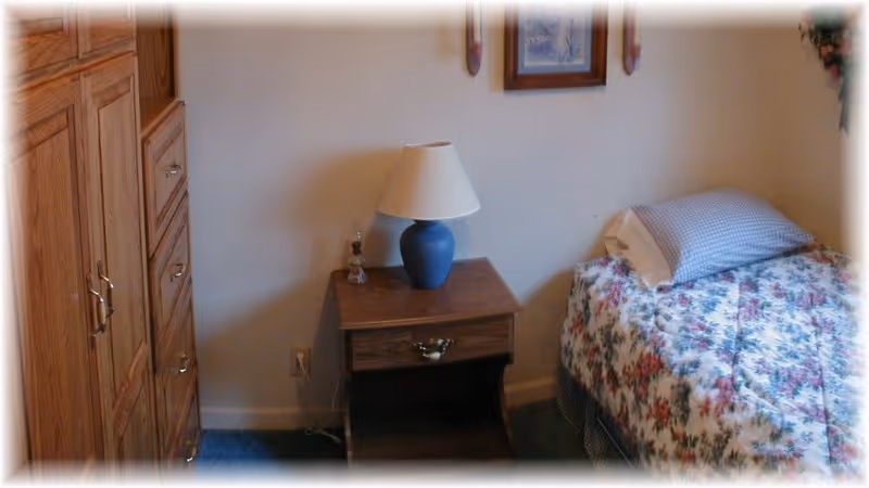 A small bedroom in a senior living facility with floral bedspread, nightstand, lamp, and wardrobe.