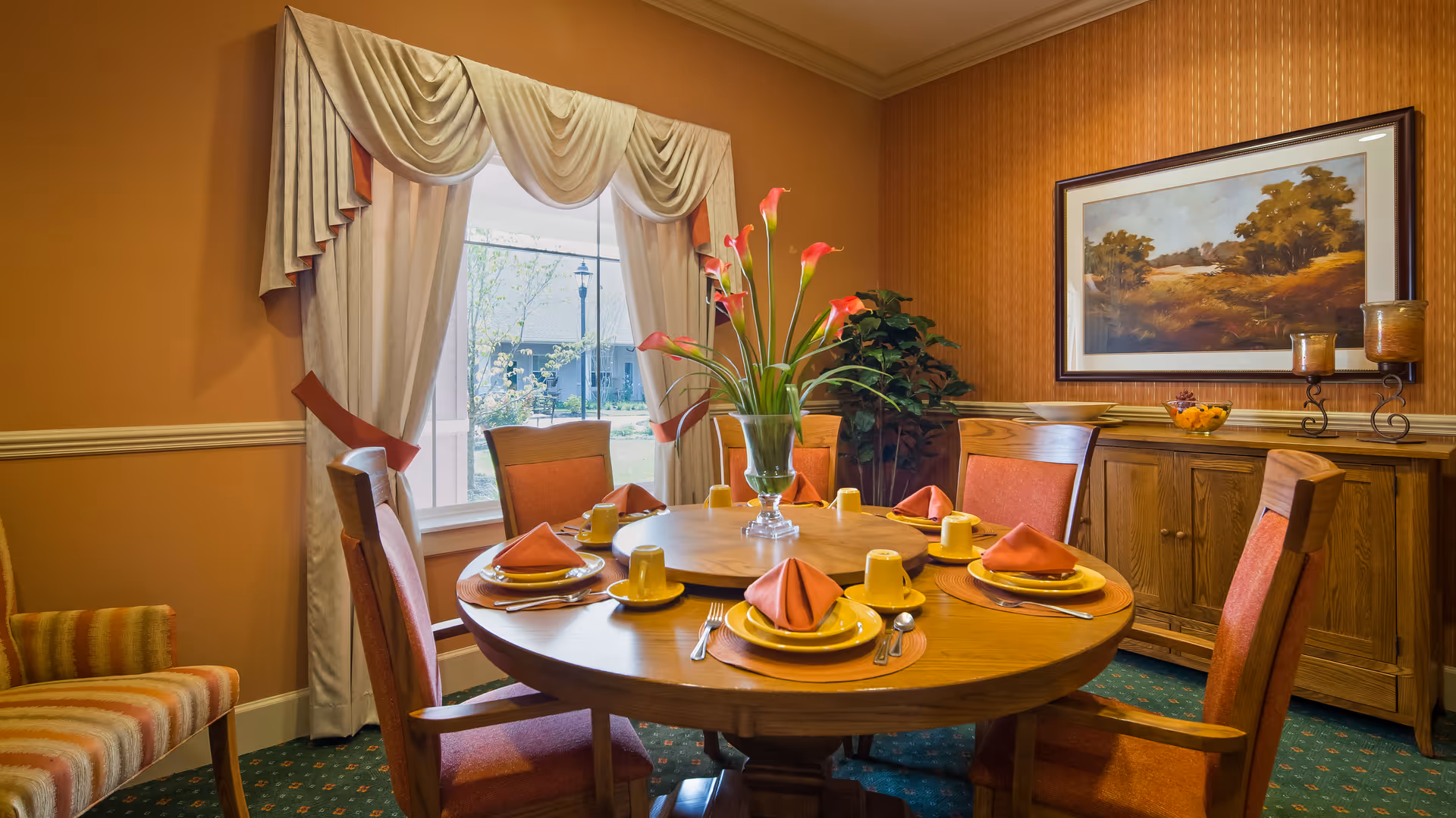 A warmly decorated dining room with a round wooden table set for six people. Each place setting includes a yellow plate, a yellow cup, silverware, and an orange folded napkin. A vase with pink flowers is centered on the table. The room features orange walls, a window with cream-colored drapes, a wooden sideboard with decorative items, and a framed landscape painting on the wall.