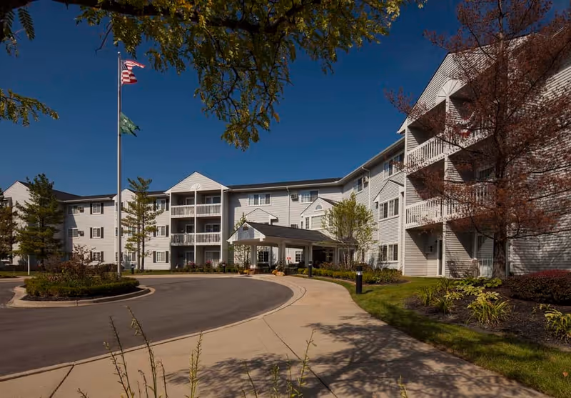 Front exterior of Waltonwood Carriage Park senior living building with a circular driveway, covered entrance, balconies, flagpole, and landscaped grounds under a clear blue sky.