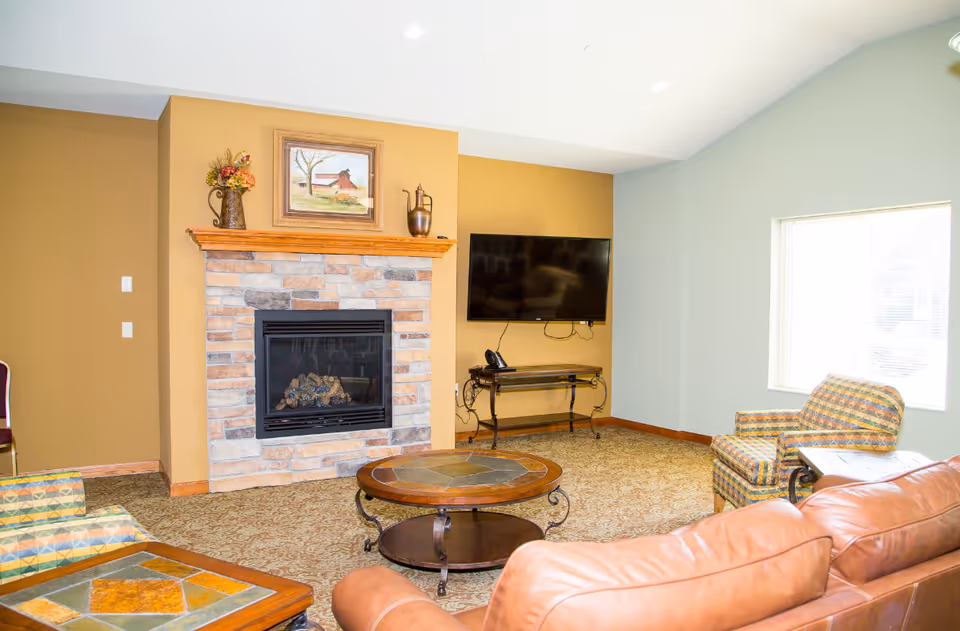 Cozy senior living lounge with stone fireplace, coffee table, leather sofa, and mounted TV.