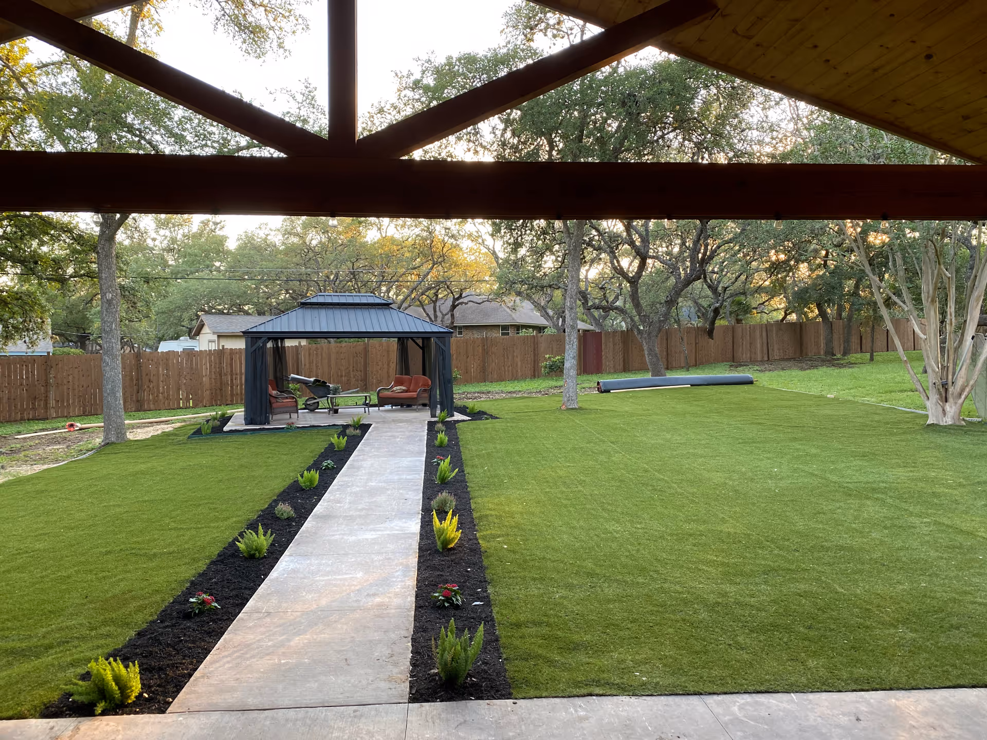 View of a well-maintained outdoor garden area with a concrete pathway leading to a gazebo furnished with chairs and a table. The garden is surrounded by a wooden fence and mature trees, with green grass covering the ground.