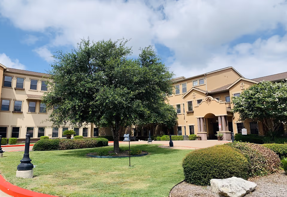 Front exterior of a beige multi-story senior living building with a large tree and landscaped lawn in the foreground.