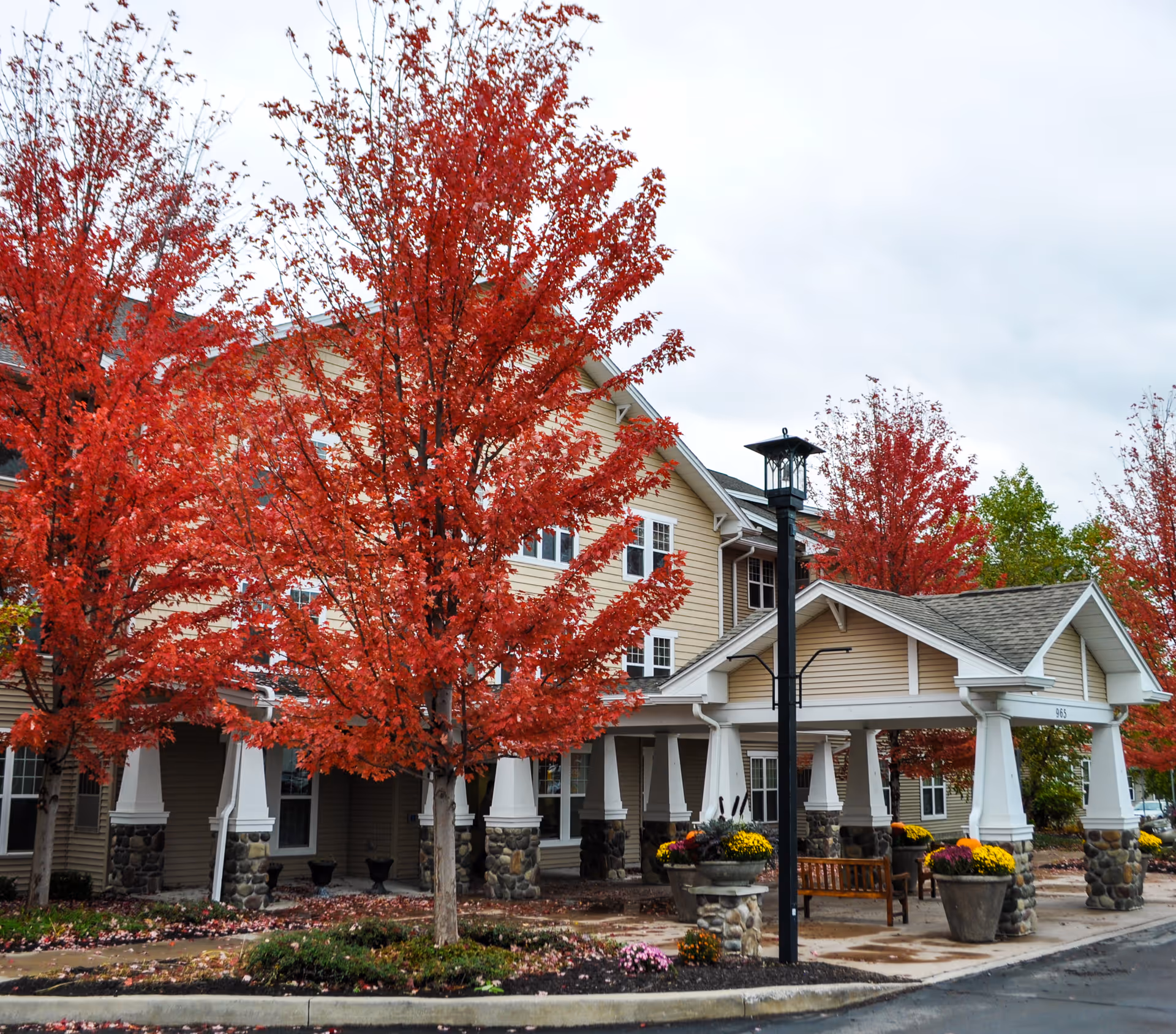 Front entrance of a multi-story senior living building with a covered porte-cochere and bright red autumn trees.