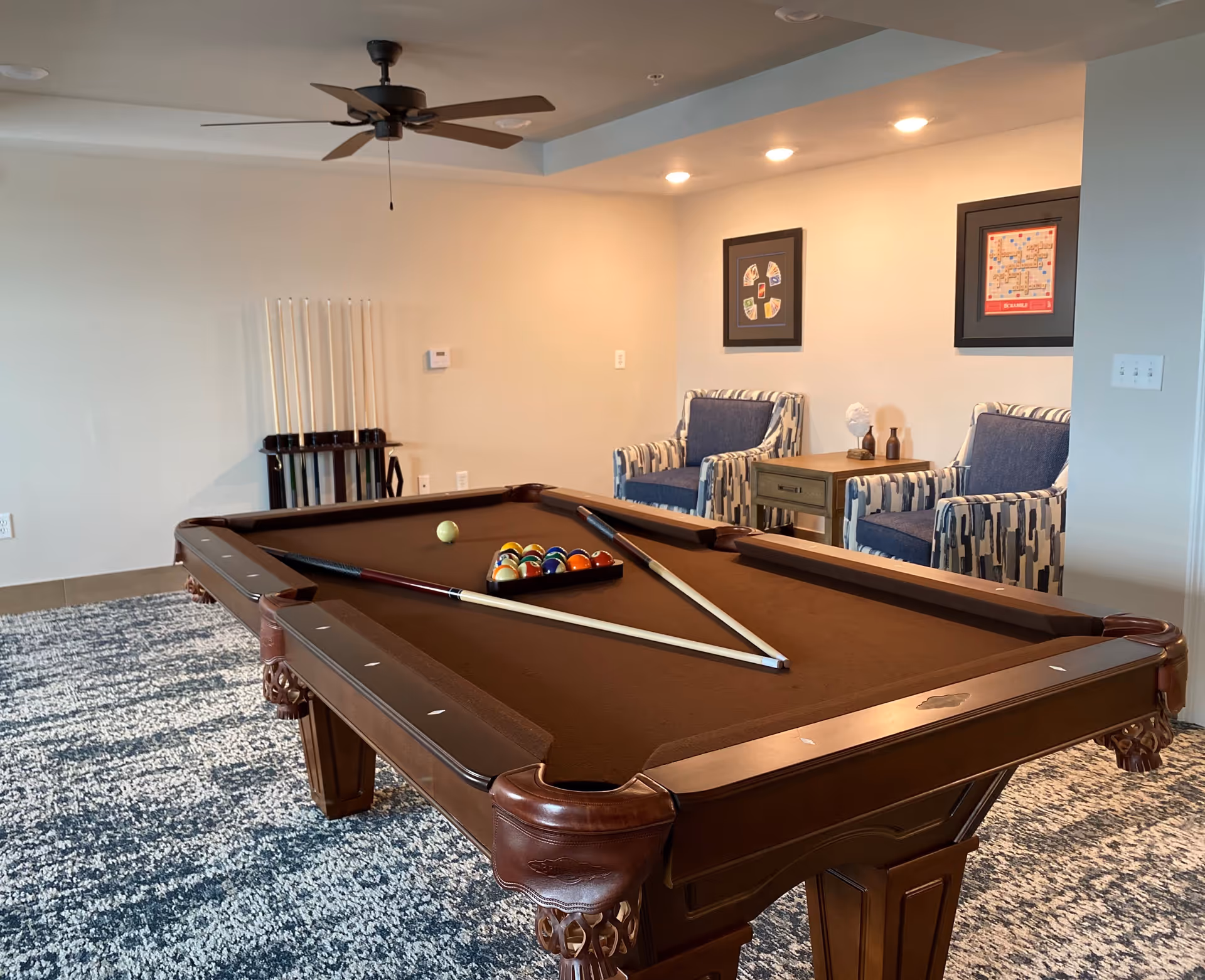 A recreational room featuring a brown pool table with pool balls arranged in a triangle and two pool cues resting on the table. Behind the pool table, there are two patterned armchairs with blue cushions and a small wooden side table between them. The room has a ceiling fan, recessed lighting, and a rack holding additional pool cues against the wall. The floor is covered with a blue and white patterned carpet.