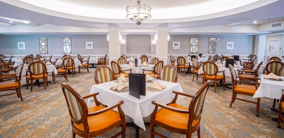 Spacious dining hall in a senior living facility with round tables and chairs.