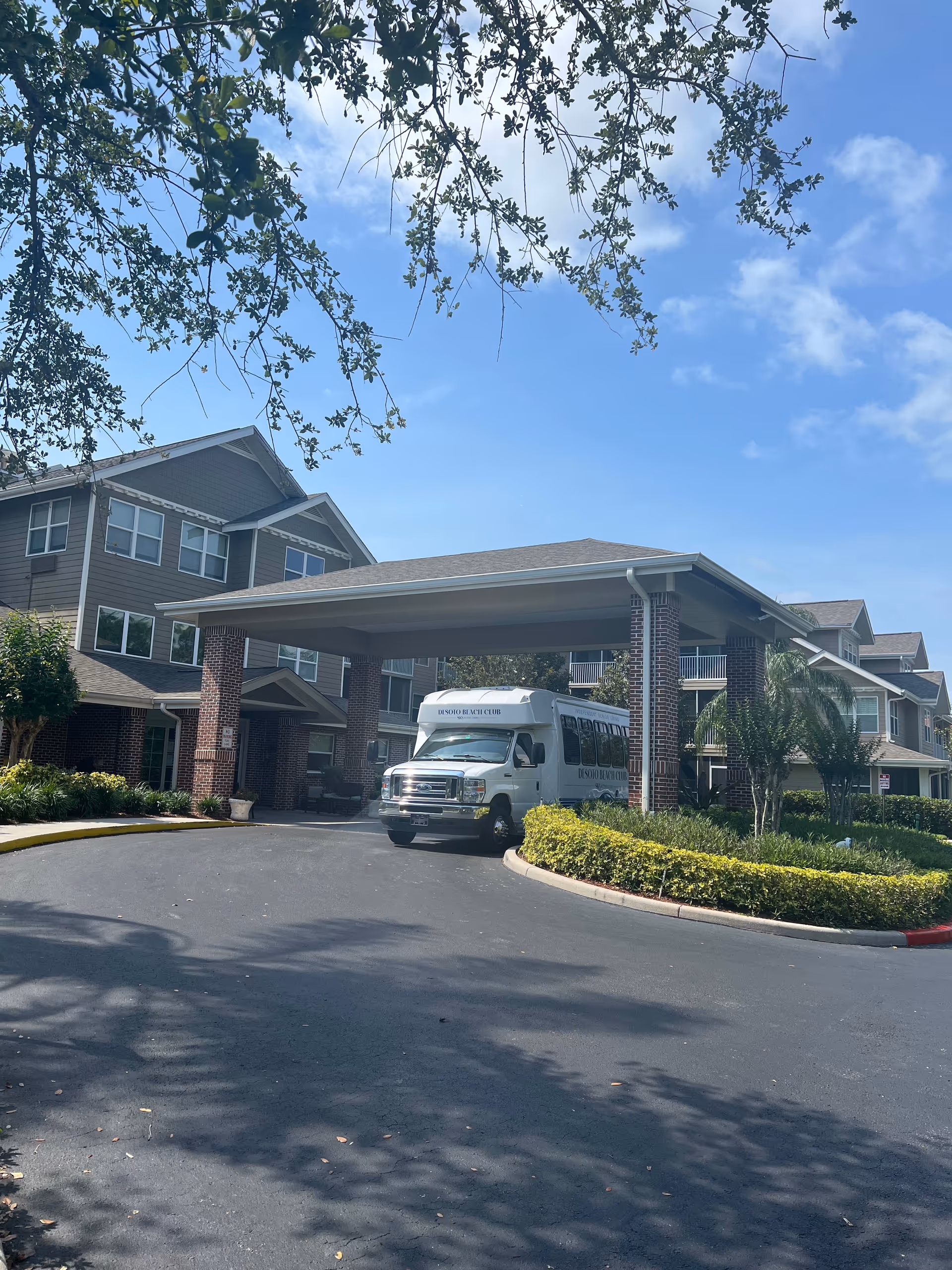 Brick senior living facility entrance with a white shuttle bus under a covered drop-off area.