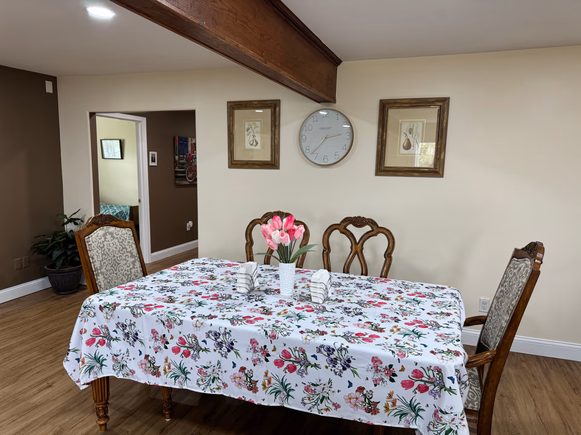 A dining room with a rectangular table covered with a floral tablecloth. The table has a vase with pink tulips and two folded napkins. There are four wooden chairs with patterned upholstery around the table. The walls are beige and brown, with two framed pictures and a clock hanging on the wall. The floor is wooden, and there is a doorway leading to another room.
