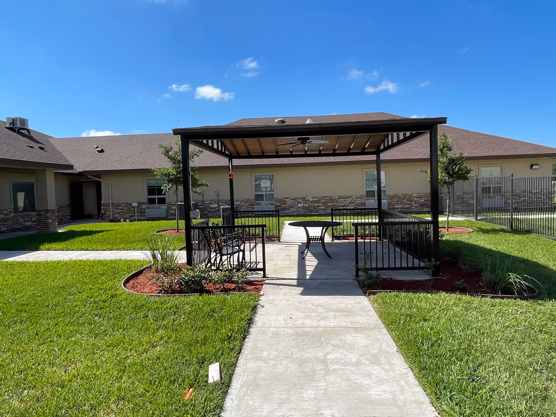 Outdoor gazebo with seating and a table in a sunny, grassy courtyard.