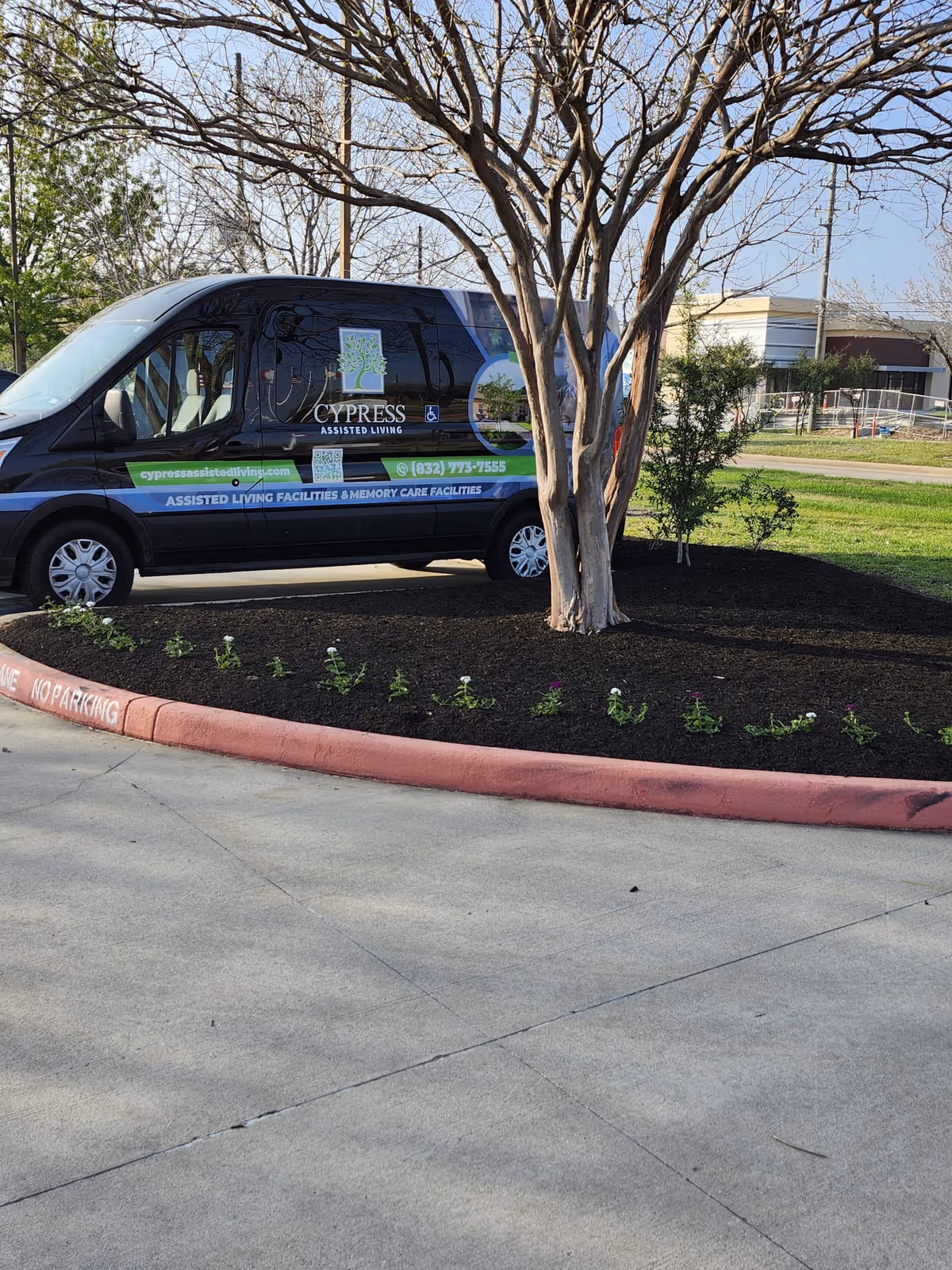 A black and blue Cypress Assisted Living van parked next to a landscaped area with a tree and small plants. The van has the Cypress Assisted Living logo, contact information, and website on its side. The surrounding area includes a concrete driveway and some greenery in the background.