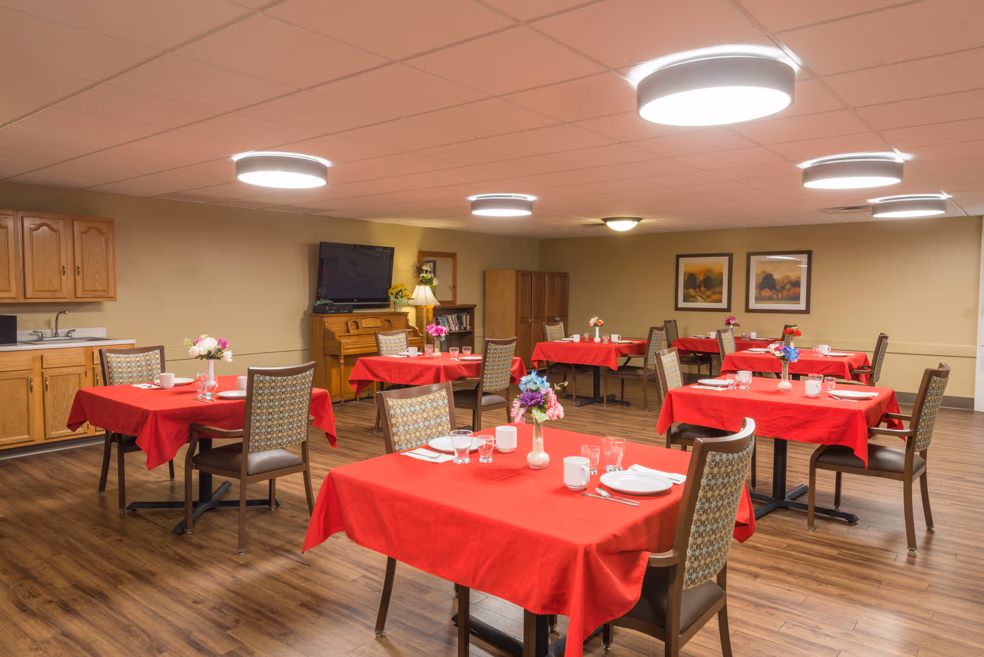 Red tablecloths and place settings fill a warm, spacious senior living dining room.