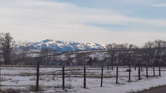Snow-covered field with fence posts and distant mountains under a cloudy sky.
