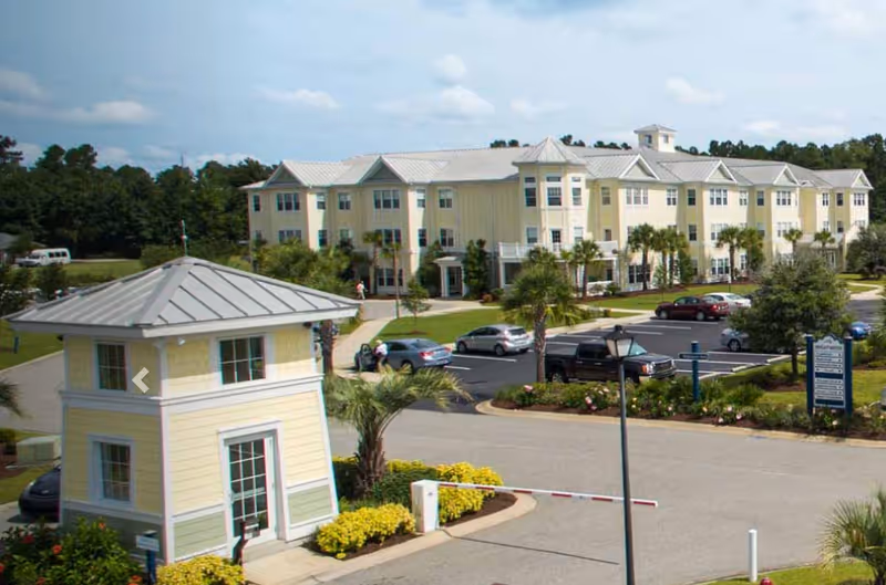 Exterior view of a senior living facility named Brightwater featuring a large, light yellow multi-story building with white trim, surrounded by landscaped greenery and palm trees. In the foreground, there is a small guardhouse with a metal roof and a parking lot with several cars parked. The sky is partly cloudy.