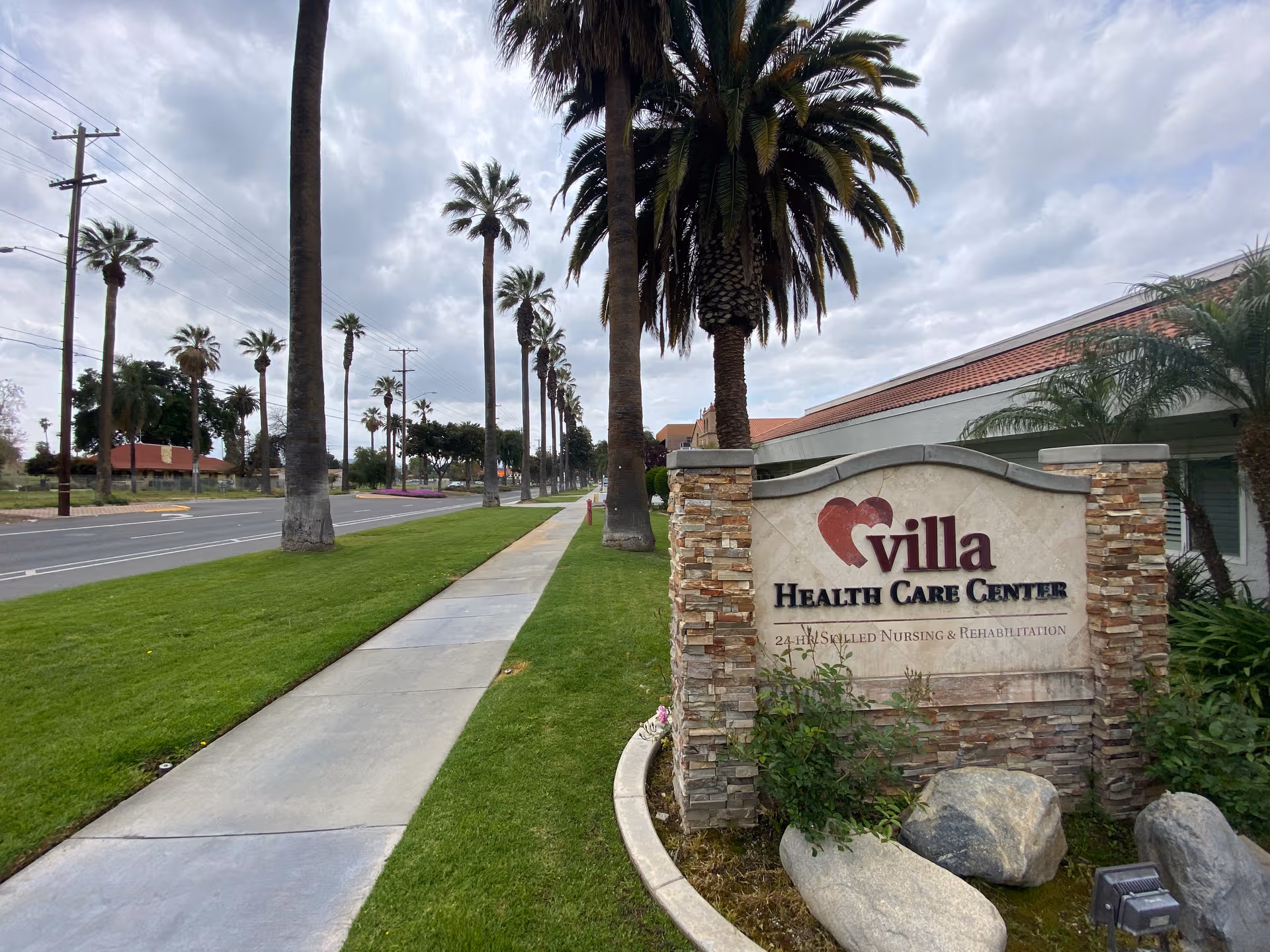 Villa Health Care Center sign beside a palm-lined road and neatly trimmed lawn.