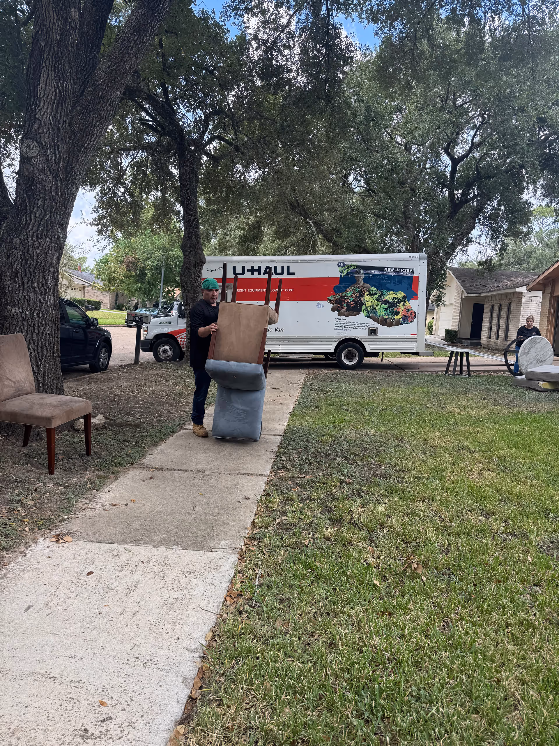 Person moving furniture outside a house with a U-Haul truck and trees nearby.