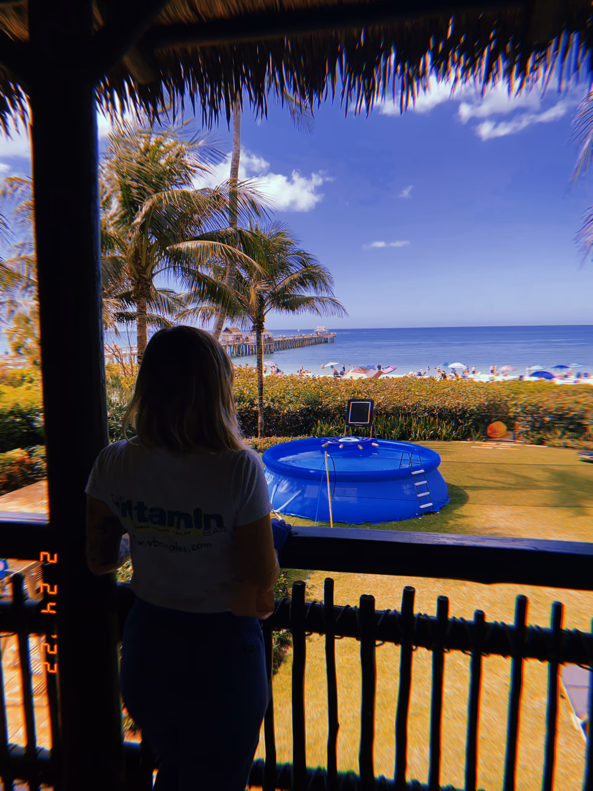 A person stands on a shaded balcony overlooking a tropical outdoor pool and beach.
